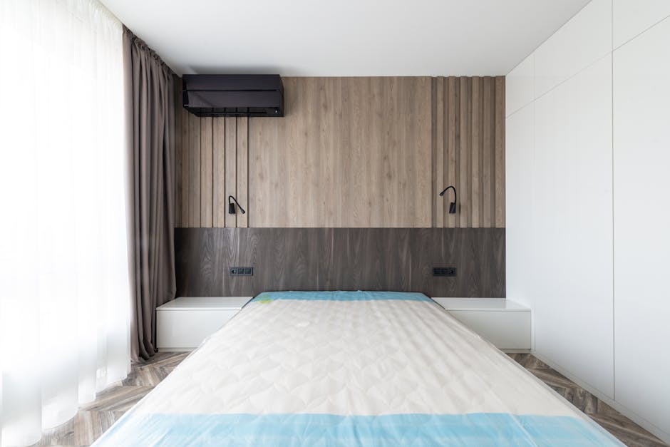 A modern bedroom featuring a white bed with a clean, textured mattress covered by a fitted sheet with light blue accents. The room has a wooden feature wall behind the bed, composed of light-colored vertical panels, with two black adjustable reading lamps mounted on the wall on either side of the bed. integrated power outlets are visible below each lamp. On the left, a sheer white curtain and a floor-length dark beige curtain frame a large window letting in natural light, illuminating the room's neat and hygienic appearance. The ceiling and adjacent walls are white, and the flooring consists of patterned wood-style laminate. Overall, the room appears well-maintained and tidy, emphasizing cleanliness and surface hygiene, suitable for domestic surface cleaning and sanitisation as promoted by Carpet Cleaners Kentish Town.