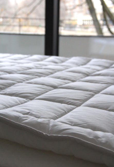 Close-up of a clean, white mattress with a quilted surface in a well-lit bedroom. The mattress appears to be freshly sanitised and free of dust or stains, with smooth, neatly stitched fabric. In the background, the room features large windows allowing natural light to illuminate the space, highlighting the modern decor. The scene emphasizes surface cleaning and deep cleaning services provided by Carpet Cleaners Kentish Town, focusing on maintaining hygiene and cleanliness of domestic bedding surfaces to promote healthy sleeping environments.