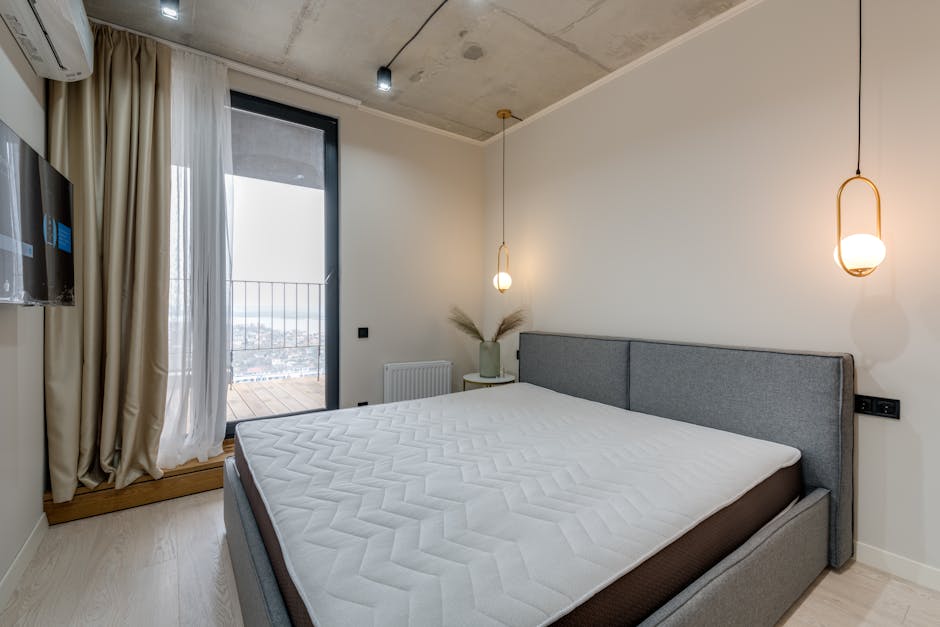 A modern bedroom featuring a white bed with a clean, textured mattress covered by a fitted sheet with light blue accents. The room has a wooden feature wall behind the bed, composed of light-colored vertical panels, with two black adjustable reading lamps mounted on the wall on either side of the bed. integrated power outlets are visible below each lamp. On the left, a sheer white curtain and a floor-length dark beige curtain frame a large window letting in natural light, illuminating the room's neat and hygienic appearance. The ceiling and adjacent walls are white, and the flooring consists of patterned wood-style laminate. Overall, the room appears well-maintained and tidy, emphasizing cleanliness and surface hygiene, suitable for domestic surface cleaning and sanitisation as promoted by Carpet Cleaners Kentish Town.