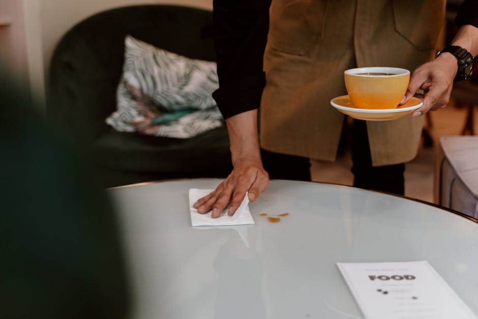 A person dressed in a brown apron is cleaning a white, glossy round table surface with a white cloth, wiping away a small spill. They are holding a beige saucer with an orange cup of tea or coffee in 