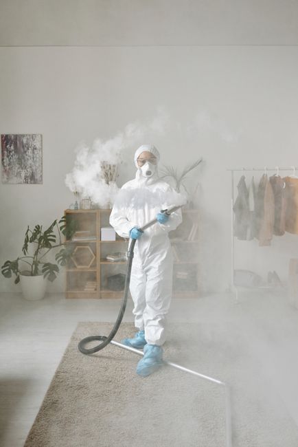 A person dressed in full protective gear, including a white coverall suit, face mask, gloves, and shoe covers, is using a fogging device to disinfect a light-colored carpet in a well-lit, modern livin