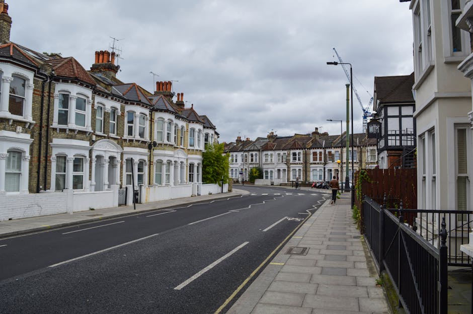 A residential street in Kentish Town with rows of Victorian terraced houses featuring bay windows, brick and white-painted facades, and pitched roofs with chimneys under a cloudy sky. The paved sidewalk runs alongside the black metal fences and gate entrance to some properties, with a few pedestrians walking along the street. The scene is well-lit with natural daylight, and the street appears clean and orderly, exemplifying typical domestic urban architecture. This image relates to surface cleaning and maintenance in residential areas, as highlighted on the website carpetcleanerskentishtown.co.uk, under the page for emergency same-day cleaning services in Kentish Town Road N19, provided by Carpet Cleaners Kentish Town.