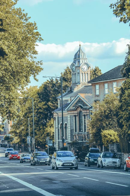 A view of a residential street on Kentish Town Road N19 during daytime, featuring a historic church with a clock tower and a pointed spire in the background. The street is lined with mature trees providing shade, and several parked and moving cars are visible along the curb. The houses have brick and stucco facades, with some windows reflecting sunlight. The sky is clear with a few scattered clouds, and the scene is well-lit, highlighting the cleanliness and orderliness of the area, emblematic of professional surface cleaning and maintenance services offered by Carpet Cleaners Kentish Town.