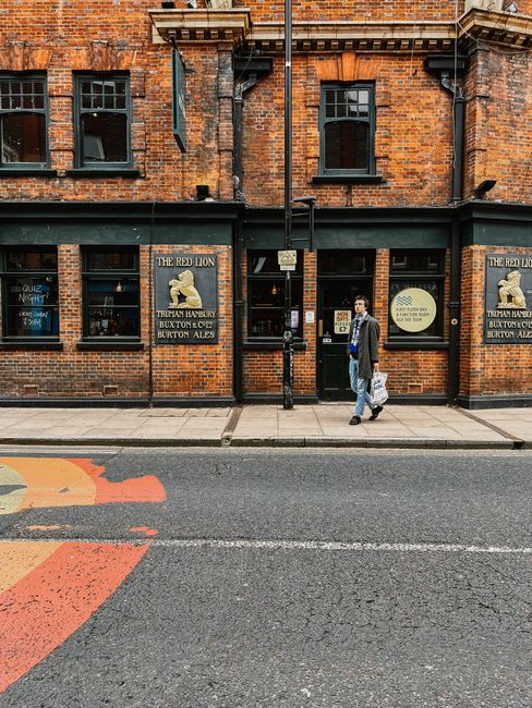 A narrow outdoor street scene outside a pub on Kentish Town Road, featuring a brick building with large glass windows, colorful signage for 'California Burrito Bar,' and hanging potted plants on the b