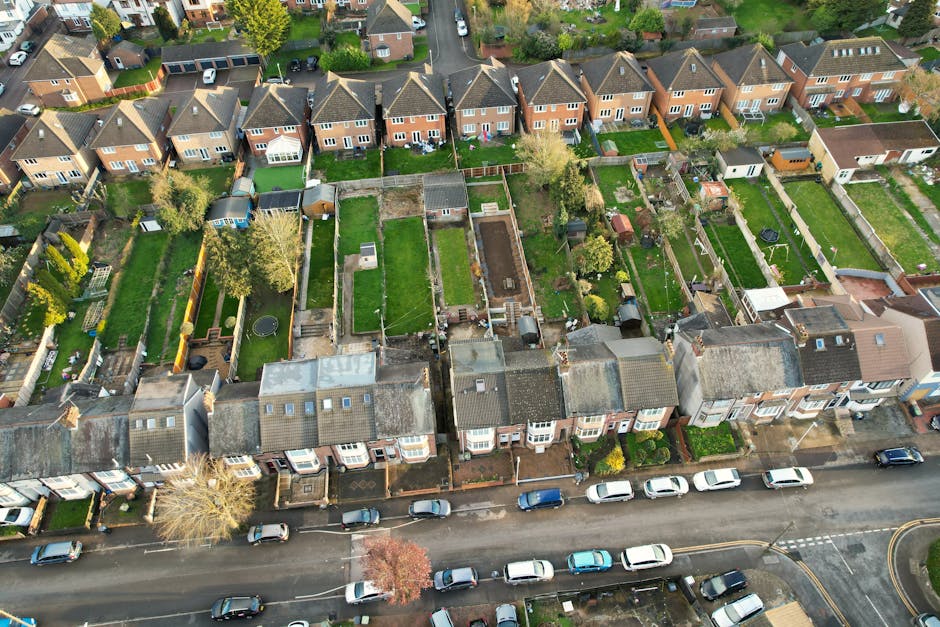 An aerial view of a residential street in Kentish Town showing closely spaced terraced houses with pitched roofs, front gardens, and parked cars along the curb. Behind the houses are backyards with various features such as lawns, small trees, patios, fences, and gardening tools. The street is lined with trees and has a row of parked vehicles. The lighting appears natural, indicating a clear day, and the scene emphasizes well-maintained outdoor spaces. The image highlights neighborhood cleanliness and the orderly appearance of the homes, consistent with professional cleaning and maintenance standards that Carpet Cleaners Kentish Town might promote in their surface cleaning and domestic cleaning services.