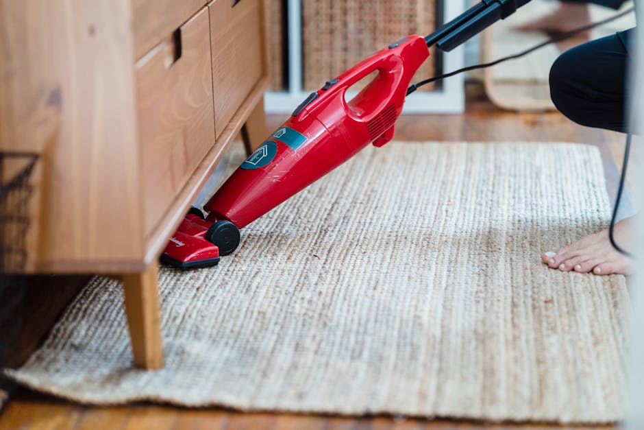 A professional cleaning technician dressed in full protective gear, including a white coverall suit, face mask, and blue gloves, is performing deep carpet cleaning in a bright, modern living room. The technician is using a steam or extraction machine connected to a black hose to thoroughly clean a plush, beige area rug positioned on light-colored wooden flooring. The room features natural light streaming through large windows, with a stylish wooden shelving unit on the right side holding books, decorative objects, and framed photos. Potted green plants and minimalistic decor enhance the clean and contemporary aesthetic of the space. The scene emphasizes surface cleaning and maintenance, exemplifying the hygiene practices of Carpet Cleaners Kentish Town, a reputable company specializing in domestic cleaning services.