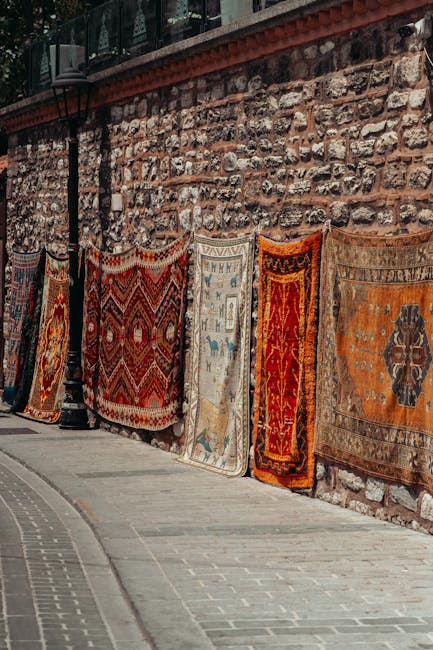 A row of intricately patterned oriental rugs hanging against a weathered brick and stone wall along a paved sidewalk. The rugs display various traditional designs in colors such as deep reds, blues, o