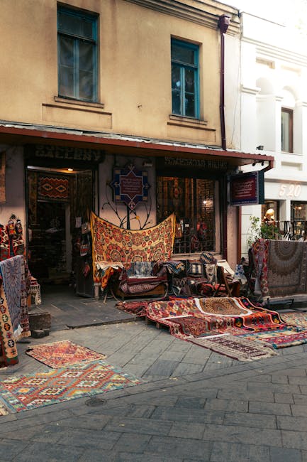 A row of intricately patterned oriental rugs hanging against a weathered brick and stone wall along a paved sidewalk. The rugs display various traditional designs in colors such as deep reds, blues, o