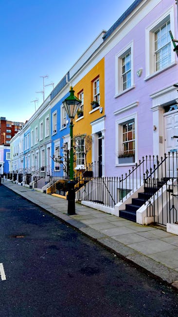 A row of colorful terraced houses in Kentish Town N19, featuring pastel-colored facades in shades of yellow, purple, light blue, and white. Each house has a small stoop with black metal railings, white-framed sash windows, and traditional door designs. A black streetlamp stands along the sidewalk, which is paved with light grey stone slabs. The sky is clear and blue, indicating a bright, sunny day. The scene is well-maintained, with clean pavements and neatly painted exteriors, representing a vibrant residential area. Carpet Cleaners Kentish Town's presence is noted for providing surface cleaning and domestic deep cleaning services, ensuring these homes remain hygienic and well-kept.