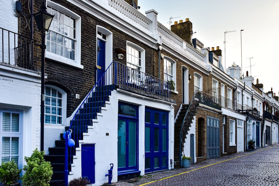 A row of multi-story residential buildings along a cobblestone street, featuring brick facades with white trim and large glass windows. The buildings have various exterior staircase designs with blue railings, leading to upper-floor entrances. Some properties have small balconies with metal railings, and potted plants can be seen on a few balconies and windowsills. The street is clean and illuminated by natural daylight, highlighting the neat, well-maintained appearance of the facades and surroundings. The image showcases a typical residential street scene in Kentish Town, emphasizing exterior architecture and street-level details that may require regular surface cleaning and maintenance, as part of professional property hygiene services by Carpet Cleaners Kentish Town.
