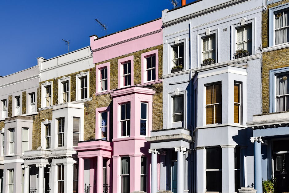A row of colorful terraced houses in Kentish Town N19, featuring pastel-colored facades in shades of yellow, purple, light blue, and white. Each house has a small stoop with black metal railings, white-framed sash windows, and traditional door designs. A black streetlamp stands along the sidewalk, which is paved with light grey stone slabs. The sky is clear and blue, indicating a bright, sunny day. The scene is well-maintained, with clean pavements and neatly painted exteriors, representing a vibrant residential area. Carpet Cleaners Kentish Town's presence is noted for providing surface cleaning and domestic deep cleaning services, ensuring these homes remain hygienic and well-kept.